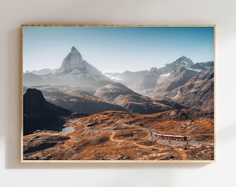 May include: A framed print of a mountain landscape featuring the Matterhorn. The image shows a train winding through a valley with brown and orange terrain. The sky is a clear blue, and the mountain peaks are covered in snow.