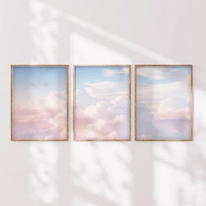 May include: Three framed prints of a pastel pink and blue sky with fluffy white clouds. The frames are light brown wood.