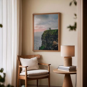 May include: A framed landscape photograph of a green cliffside overlooking the ocean at sunset, displayed on a wall above a wooden armchair with a cream cushion, next to a small round wooden table holding a lamp and a stack of books.