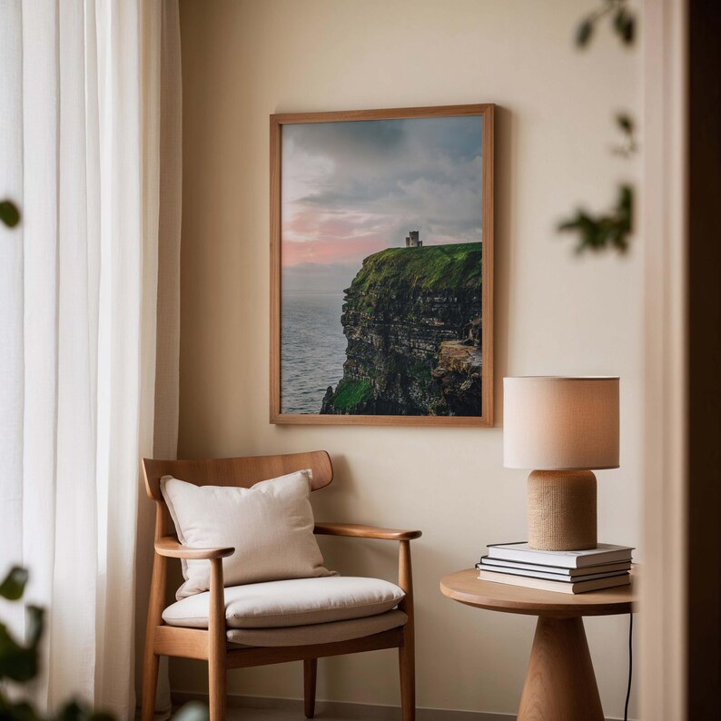May include: A framed landscape photograph of a green cliffside overlooking the ocean at sunset, displayed on a wall above a wooden armchair with a cream cushion, next to a small round wooden table holding a lamp and a stack of books.