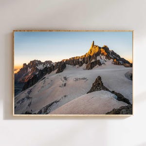 Może przedstawiać: Oprawiona fotografia krajobrazu ośnieżonego pasma górskiego o wschodzie słońca. Szczyty oświetlone są złotym światłem, kontrastującym z błękitnym niebem. Rama wykonana jest z jasnego drewna.