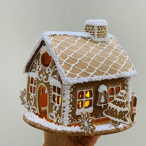 Può includere: Una casetta di pan di zenzero decorata con glassa bianca e una finestra illuminata. La casa ha un tetto marrone con un camino bianco e una base ricoperta di neve bianca.