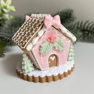 Puede incluir: Una casa de jengibre rosa y marrón con glaseado blanco y un lazo en la parte superior. La casa está decorada con hojas de acebo verdes y bayas rojas. La casa está sentada sobre una base de galleta marrón y blanca.