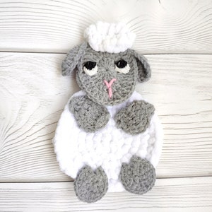 Może przedstawiać: Pluszowa zabawka owca wykonana na szydełku w kolorze szarym i białym z białą, puszystą głową i czarnymi oczami. Owca siedzi na białym drewnianym podłożu.