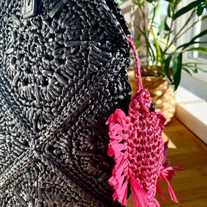 May include: Black woven raffia tote bag with a textured surface. A bright pink fish-shaped charm hangs from the bag. The charm has a fringed tail and fins. The bag is partially visible.