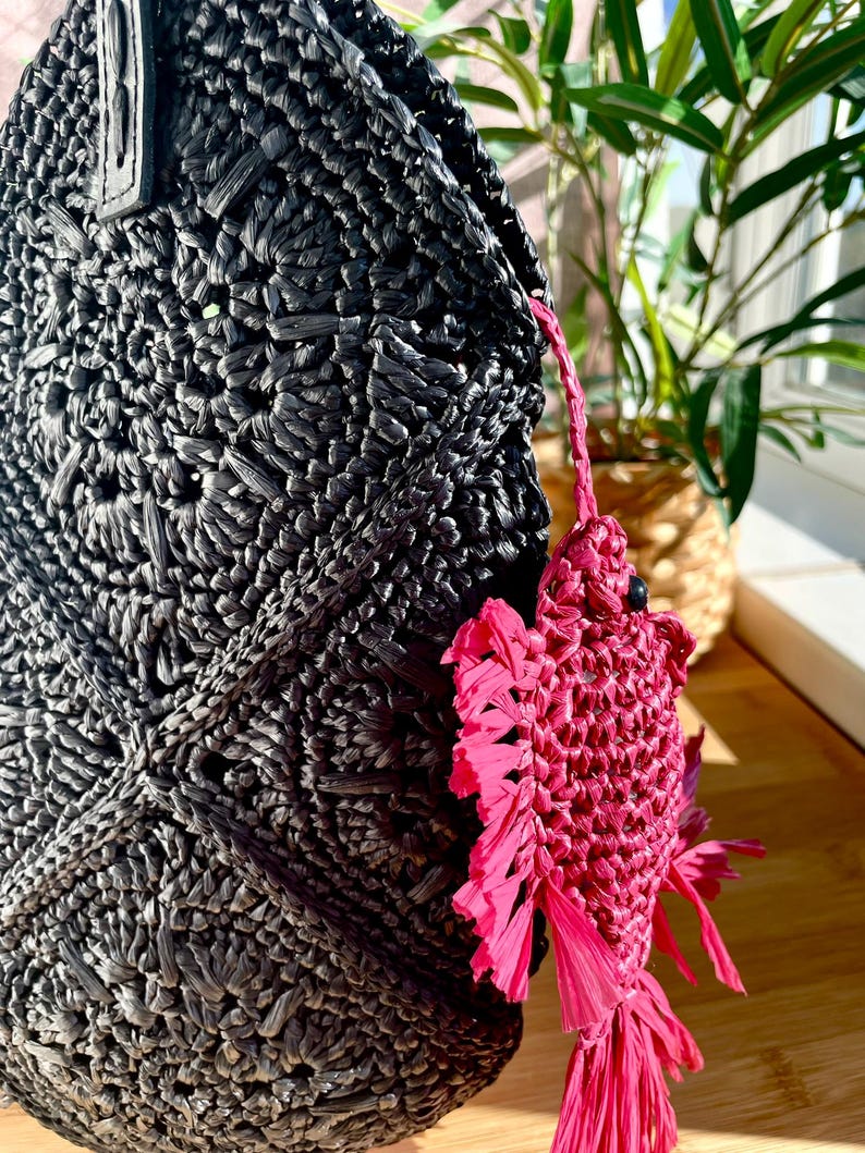 May include: Black woven raffia tote bag with a textured surface. A bright pink fish-shaped charm hangs from the bag. The charm has a fringed tail and fins. The bag is partially visible.