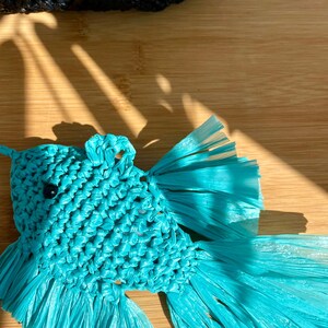 May include: A handcrafted, turquoise fish-shaped ornament. The fish is made from woven material, with a detailed body and flowing tail fins. A small black bead serves as the eye. The background includes a wooden surface and a portion of a black woven item.