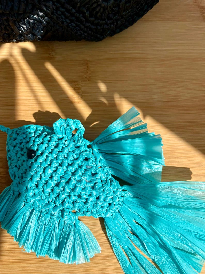May include: A handcrafted, turquoise fish-shaped ornament. The fish is made from woven material, with a detailed body and flowing tail fins. A small black bead serves as the eye. The background includes a wooden surface and a portion of a black woven item.