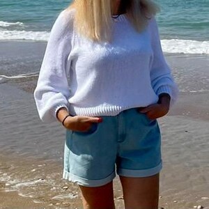 K&ouml;nnte beinhalten: Ein wei&szlig;er, lang&auml;rmeliger Strickpullover mit Rundhalsausschnitt und gerippten B&uuml;ndchen, kombiniert mit hellblauen Jeansshorts mit umgeschlagenem Saum. Der Pullover hat eine lockere Passform mit leicht bauschigen &Auml;rmeln und die Shorts.