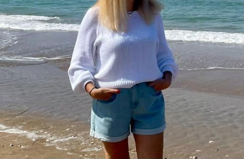 K&ouml;nnte beinhalten: Ein wei&szlig;er, lang&auml;rmeliger Strickpullover mit Rundhalsausschnitt und gerippten B&uuml;ndchen, kombiniert mit hellblauen Jeansshorts mit umgeschlagenem Saum. Der Pullover hat eine lockere Passform mit leicht bauschigen &Auml;rmeln und die Shorts.