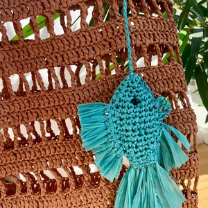 May include: A brown woven tote bag with a turquoise fish-shaped keychain. The bag has a large, open weave design. The fish keychain is made of woven material with fringed fins and tail. The background includes green foliage.