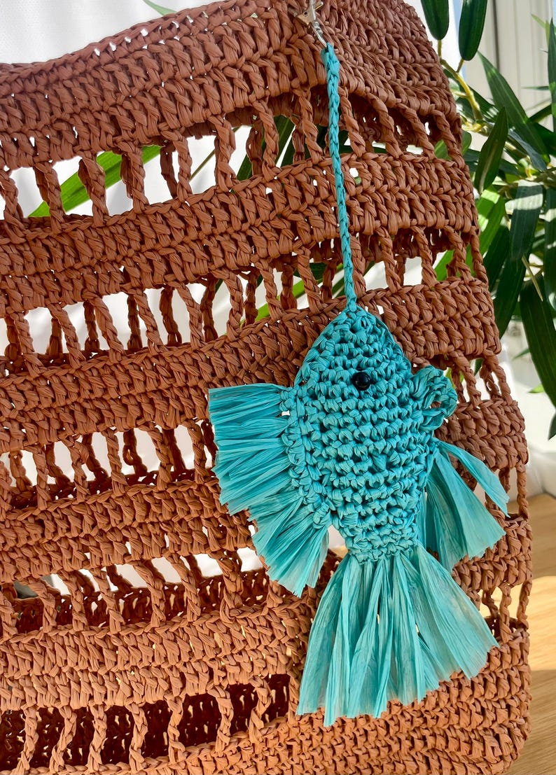 May include: A brown woven tote bag with a turquoise fish-shaped keychain. The bag has a large, open weave design. The fish keychain is made of woven material with fringed fins and tail. The background includes green foliage.