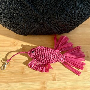 May include: A handcrafted, magenta-colored raffia fish keychain with a silver-tone clasp. The fish has a textured body and fringed fins. A black woven bag is in the background, resting on a wooden surface.