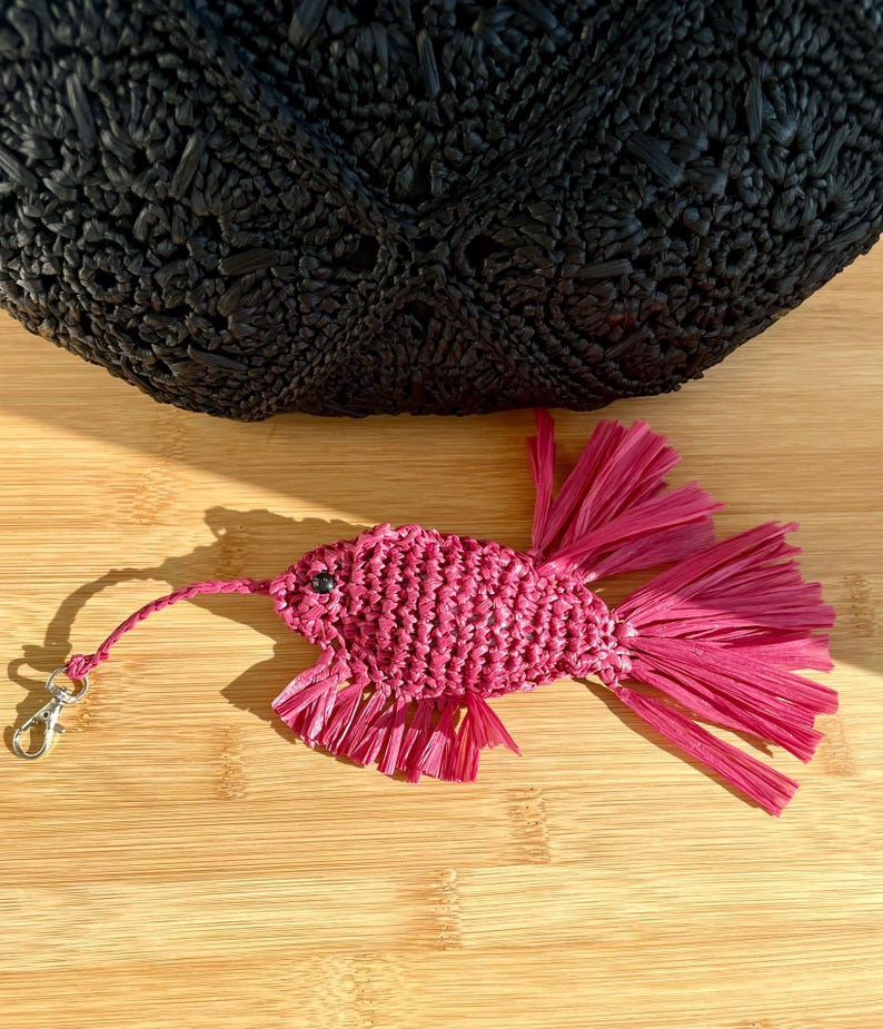 May include: A handcrafted, magenta-colored raffia fish keychain with a silver-tone clasp. The fish has a textured body and fringed fins. A black woven bag is in the background, resting on a wooden surface.