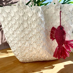 May include: Two white woven tote bags with handles, featuring a textured pattern. A small, red fish-shaped charm hangs from one bag. The bags are on a wooden surface, with sunlight casting shadows.
