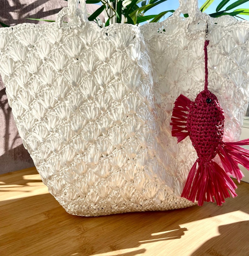 May include: Two white woven tote bags with handles, featuring a textured pattern. A small, red fish-shaped charm hangs from one bag. The bags are on a wooden surface, with sunlight casting shadows.