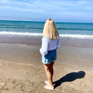 K&ouml;nnte beinhalten: Ein wei&szlig;er Pullover mit langen &Auml;rmeln und Rundhalsausschnitt sowie hellblaue Jeansshorts mit umgeschlagenem Saum. Das Outfit wird mit einfachen wei&szlig;en Zehentrennern kombiniert. Die Kleidung wird von einer Person am Sandstrand getragen.