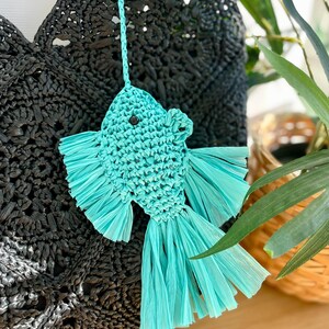 May include: A turquoise raffia fish ornament with a black button eye and fringed fins hangs in front of a black woven bag. The fish is suspended by a matching turquoise cord. Green foliage is visible in the background.