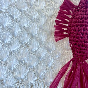 May include: Close-up of a white, textured, woven material with a repeating pattern. A magenta-colored, woven decorative element with fringe is on the right side of the image. The woven material has a fish scale-like pattern.