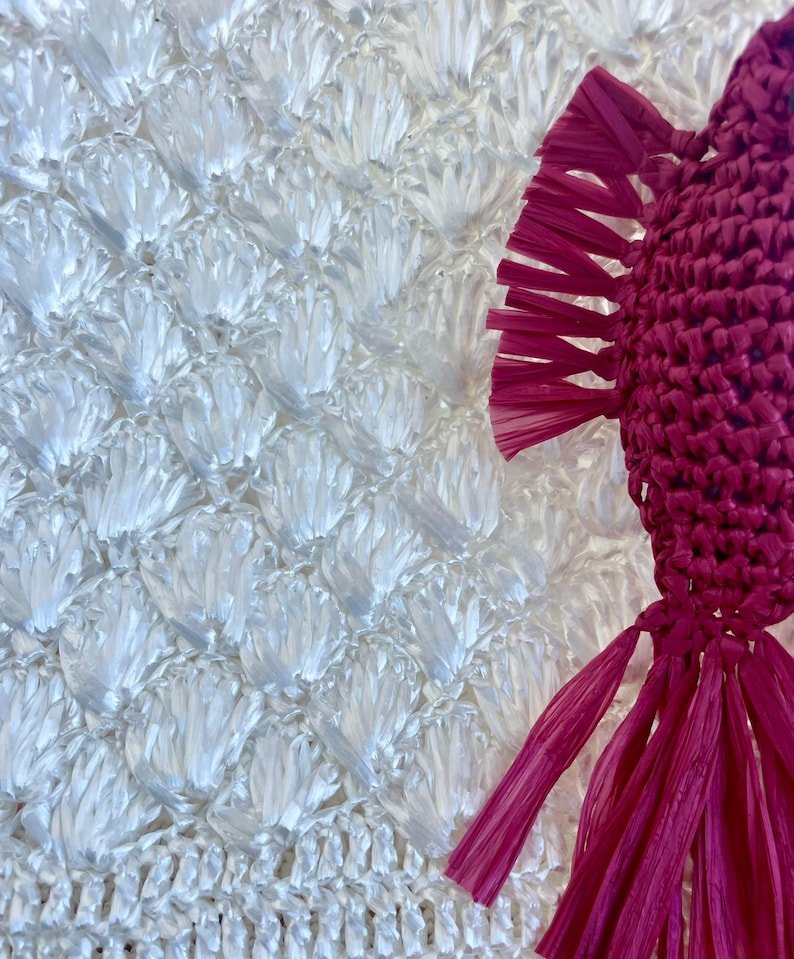 May include: Close-up of a white, textured, woven material with a repeating pattern. A magenta-colored, woven decorative element with fringe is on the right side of the image. The woven material has a fish scale-like pattern.
