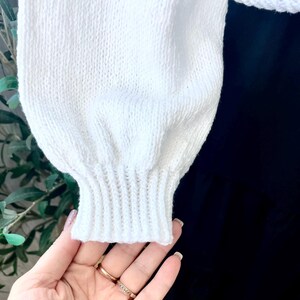 K&ouml;nnte beinhalten: Ein wei&szlig;er, lang&auml;rmeliger Strickpullover mit strukturiertem Strickmuster, volumin&ouml;sen Puff&auml;rmeln und einem gerippten B&uuml;ndchen am Handgelenk. Das Kleidungsst&uuml;ck ist mit einem einfarbigen schwarzen Rock kombiniert, was einen starken Kontrast erzeugt.
