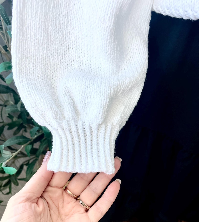 K&ouml;nnte beinhalten: Ein wei&szlig;er, lang&auml;rmeliger Strickpullover mit strukturiertem Strickmuster, volumin&ouml;sen Puff&auml;rmeln und einem gerippten B&uuml;ndchen am Handgelenk. Das Kleidungsst&uuml;ck ist mit einem einfarbigen schwarzen Rock kombiniert, was einen starken Kontrast erzeugt.