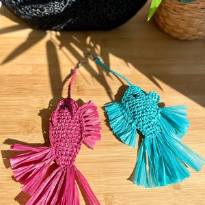 May include: Two handmade fish-shaped keychains. One is magenta, the other is turquoise. Both are made of woven raffia with fringed tails. They hang on silver-colored keyrings. The keychains are on a wooden surface.