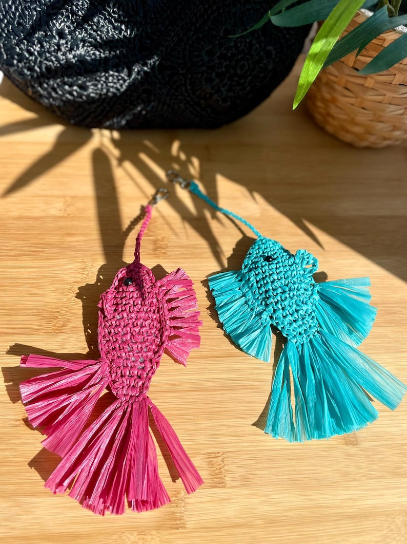 May include: Two handmade fish-shaped keychains. One is magenta, the other is turquoise. Both are made of woven raffia with fringed tails. They hang on silver-colored keyrings. The keychains are on a wooden surface.