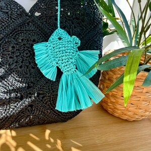 May include: A turquoise fish-shaped ornament with fringed fins and tail hangs in front of a black woven bag. The fish is made of woven material. A woven basket with green plants is in the background.