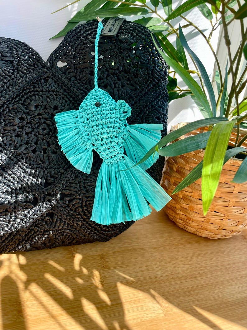 May include: A turquoise fish-shaped ornament with fringed fins and tail hangs in front of a black woven bag. The fish is made of woven material. A woven basket with green plants is in the background.