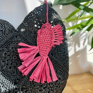 May include: A black woven tote bag with a pink fish-shaped charm. The charm is made of woven material with fringed edges. The bag has a textured surface and a round shape. The charm is attached to the bag with a black leather strap.