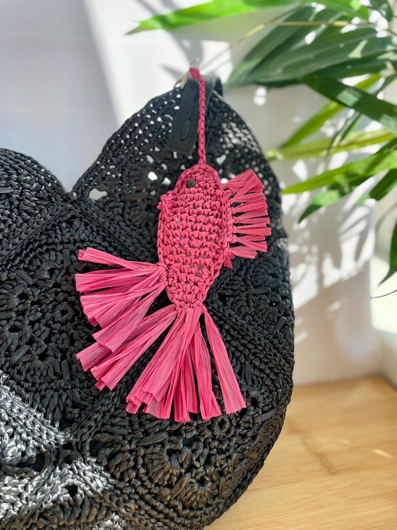 May include: A black woven tote bag with a pink fish-shaped charm. The charm is made of woven material with fringed edges. The bag has a textured surface and a round shape. The charm is attached to the bag with a black leather strap.