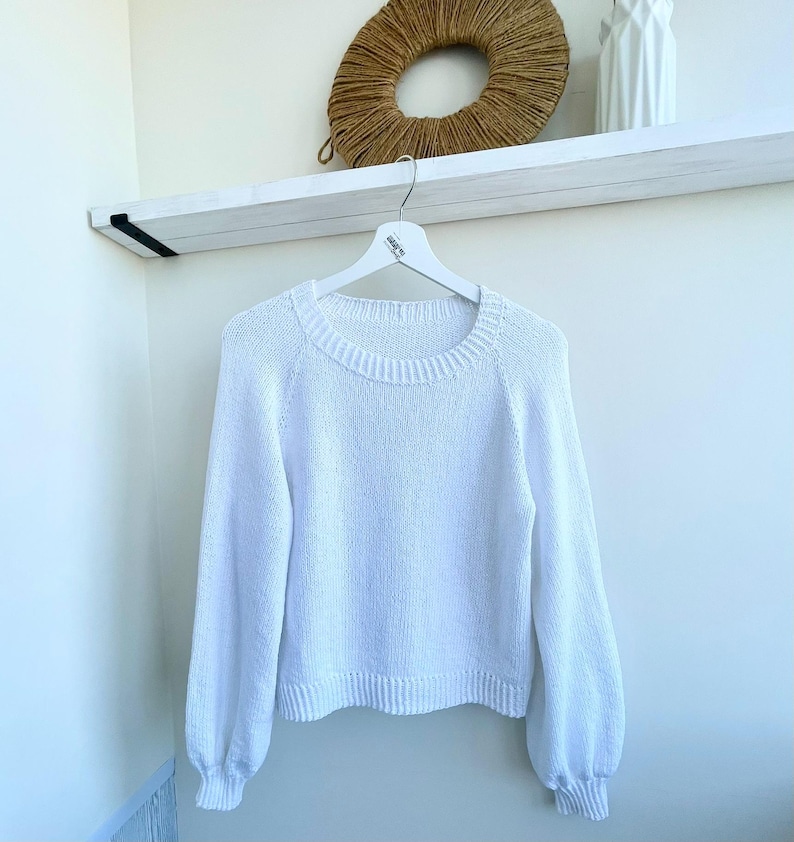 K&ouml;nnte beinhalten: Ein einfarbiger wei&szlig;er Langarmpullover mit Rundhalsausschnitt, gerippten B&uuml;ndchen und einem gerippten Saum. Das Kleidungsst&uuml;ck ist in Strickoptik gefertigt und wird auf einem wei&szlig;en Kleiderb&uuml;gel pr&auml;sentiert, was ein schlichtes Design mit &Auml;rmeln zeigt.