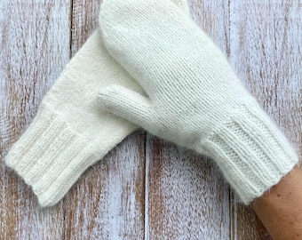 Può includere: Un paio di guanti in maglia color crema. I guanti sono realizzati in un filato morbido e strutturato e presentano polsini a coste. I guanti sono progettati per tenere le mani al caldo quando fa freddo. I guanti sono mostrati su una superficie di legno.