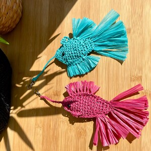May include: Two handmade fish-shaped ornaments. One is turquoise, the other is pink. Both are made of woven material with fringed tails and attached lanyards with metal clasps. They rest on a wooden surface.