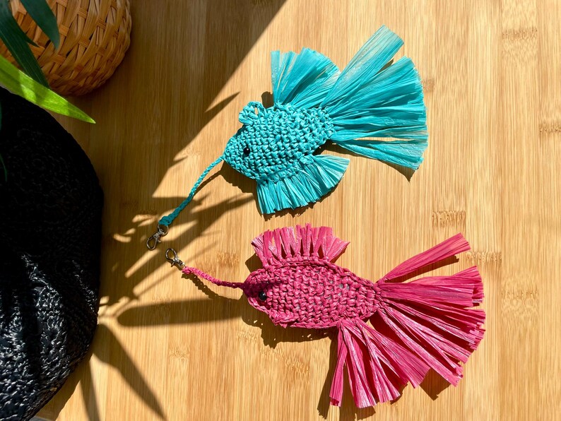 May include: Two handmade fish-shaped ornaments. One is turquoise, the other is pink. Both are made of woven material with fringed tails and attached lanyards with metal clasps. They rest on a wooden surface.