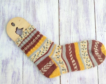 May include: A colorful knitted sock with stripes of yellow, brown, gray, and white. The sock is lying on a light purple wooden surface.