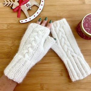 May include: A pair of off-white fingerless gloves featuring a vertical cable knit pattern and ribbed cuffs. The gloves are crafted from a soft, fuzzy yarn and extend past the wrist, providing coverage for the hands while leaving the fingers exposed.