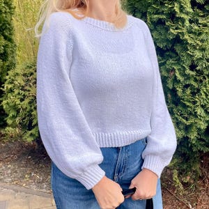 K&ouml;nnte beinhalten: Ein einfarbiger wei&szlig;er Langarmpullover mit Rundhalsausschnitt und gerippten B&uuml;ndchen an &Auml;rmeln und Saum. Das Strickkleidungsst&uuml;ck hat eine lockere Passform mit leicht bauschigen &Auml;rmeln und wird mit einer blauen Jeans kombiniert.