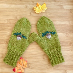 May include: A pair of lime green knit mittens featuring a small embroidered acorn design with dark green leaves and a light brown nut. The mittens have a ribbed cuff and a smooth knit texture, presented alongside two decorative autumn leaves.