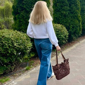 K&ouml;nnte beinhalten: Ein wei&szlig;er Langarmpullover, eine blaue Jeans und wei&szlig;e Turnschuhe, kombiniert mit einer dunkelbraunen Tragetasche mit kreisf&ouml;rmigem Ausschnittmuster und zwei lederartigen Griffen, getragen von einer Person auf einem gepflasterten Weg.