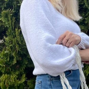 K&ouml;nnte beinhalten: Ein wei&szlig;er, lang&auml;rmeliger Strickpullover mit V-Ausschnitt und gerafften B&uuml;ndchen, kombiniert mit einer blauen Jeans. Der Pullover hat ein strukturiertes Strickmuster und eine leicht verk&uuml;rzte Passform, erg&auml;nzt durch ein wei&szlig;es geflochtenes Accessoire.
