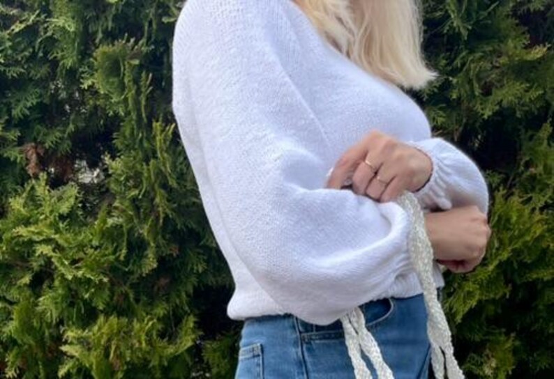 K&ouml;nnte beinhalten: Ein wei&szlig;er, lang&auml;rmeliger Strickpullover mit V-Ausschnitt und gerafften B&uuml;ndchen, kombiniert mit einer blauen Jeans. Der Pullover hat ein strukturiertes Strickmuster und eine leicht verk&uuml;rzte Passform, erg&auml;nzt durch ein wei&szlig;es geflochtenes Accessoire.