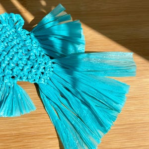 May include: A handcrafted, turquoise-colored raffia fish-shaped coaster. The coaster is made of woven raffia and has a detailed, textured design. The fish has a central body and flowing fins, perfect for protecting surfaces from drink condensation.