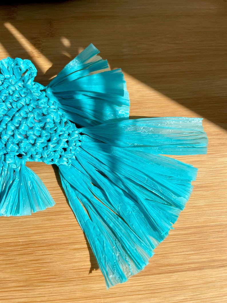 May include: A handcrafted, turquoise-colored raffia fish-shaped coaster. The coaster is made of woven raffia and has a detailed, textured design. The fish has a central body and flowing fins, perfect for protecting surfaces from drink condensation.