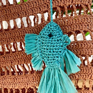 May include: A turquoise fish-shaped raffia ornament with a black eye, hanging from a woven brown bag. The fish has fringed fins and tail. The bag has a woven, open design.