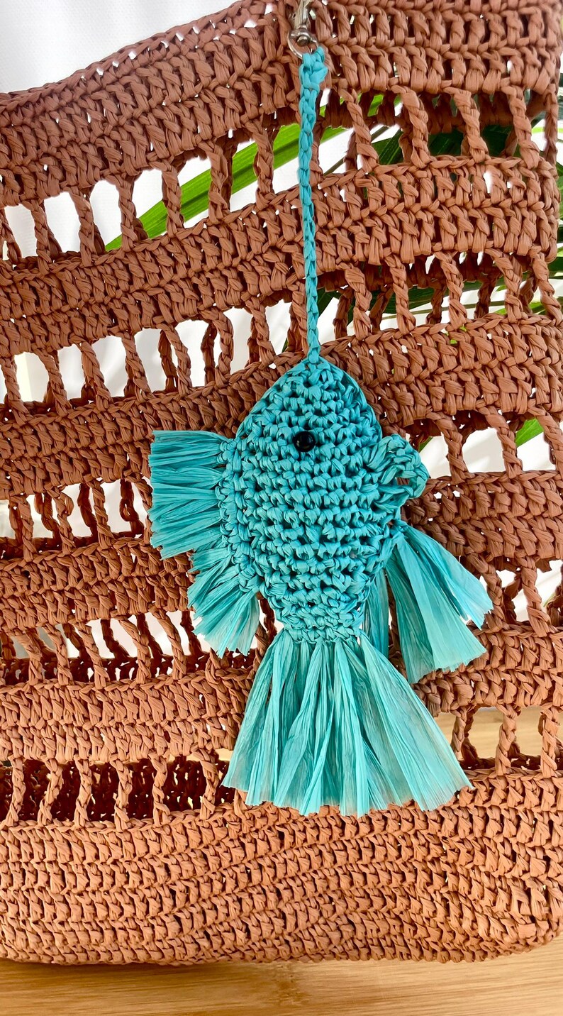 May include: A turquoise fish-shaped raffia ornament with a black eye, hanging from a woven brown bag. The fish has fringed fins and tail. The bag has a woven, open design.