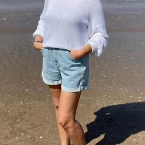 K&ouml;nnte beinhalten: Ein wei&szlig;er, lang&auml;rmeliger Strickpullover mit Rundhalsausschnitt und gerippten B&uuml;ndchen, kombiniert mit hellblauen Jeansshorts mit umgeschlagenem Saum und Seitentaschen. Der Pullover hat eine lockere Passform und die Shorts ein klassisches Design.
