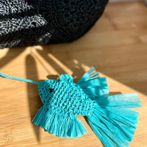 May include: A turquoise fish-shaped ornament made of woven material and fringed paper. The fish has black bead eyes and a string for hanging. A black woven bag is in the background, resting on a wooden surface.
