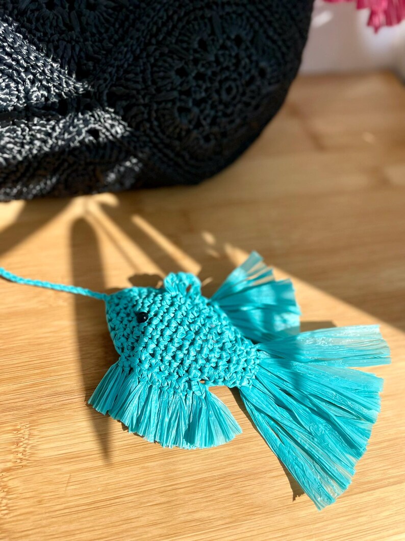 May include: A turquoise fish-shaped ornament made of woven material and fringed paper. The fish has black bead eyes and a string for hanging. A black woven bag is in the background, resting on a wooden surface.